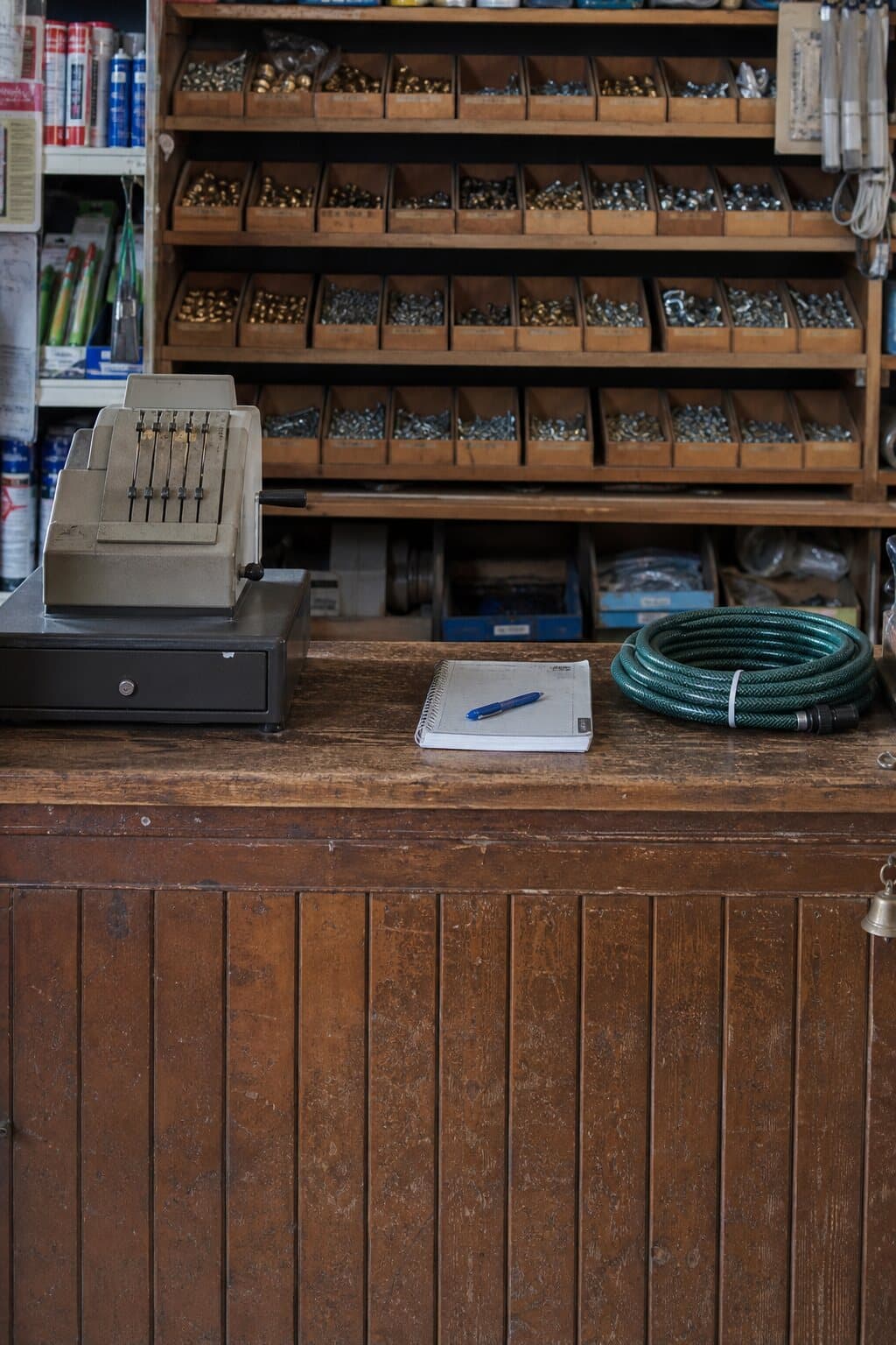 The wooden service counter of a small independent hardware shop, with an old till and labelled bins of fittings behind it.